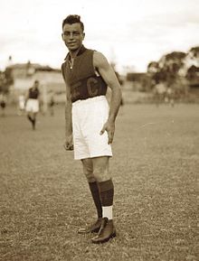 A tall, fair-skinned man looking at the camera with dark, slicked-back hair, leaning to one side. He is wearing a sleeveless, collared shirt with a red top half and blue bottom half, with "FC" in capital letters emblazoned across the front. He is wearing white shorts, knee-high socks and boots, standing against an out-of-focus background with trees and other players visible. The photograph is sepia-toned.