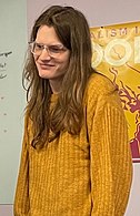 A white woman with brown eyes and long, brown hair. She is wearing clear glasses and a yellow sweater while facing slightly to the left.