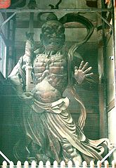 Front view of a scary statue carrying a spear like object in his right hand and the palm of his left hand facing the viewer with fingers spread. Sculpted breastplate and necklace are visible. Color photograph.