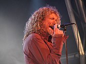 A man with long, curly hair wearing a red dress shirt and singing into a microphone on a stand.