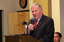 Man in black suit standing behind a podium. He is speaking into a microphone, which he is holding.