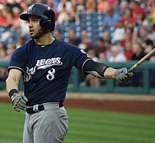 A man wearing a navy blue Brewers jersey, gray pants, and navy blue cap takes a practice swing with his bat