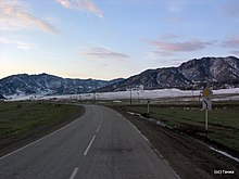 Route dans une vallée avec un virage, avec en arrière-plan des montagnes. Les hauteurs sont enneigées, mais la vallée ne l'est qu'en partie.