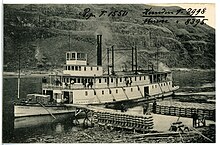 A steamboat waits at a pier on a river bank while sacks of cargo are loaded.