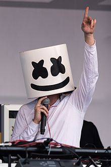 Marshmello holding a microphone in his right hand with his left hand raised up