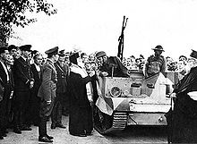 Photographie noir et blanc : un pope aux côtés d'une chenillette, sous les yeux de nombreux hommes, dont des soldats.