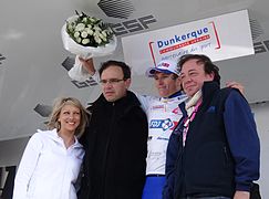 De trois-quarts, quatre personnes alignées, habillés de blanc ou de noir, sourient. Le cycliste lève un bouquet blanc.