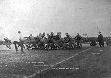 A grainy black and white photo of teams in sports jerseys and light padding contesting a play near the touchline, while watched by officials, coaches and crowds.