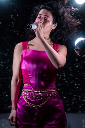 A young brunette woman wearing a purple leotard and singing into a microphone.