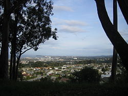 view of Millbrae, facing southeast from Junipero Serra Park, with SFO runways, the Westin SFO, and Millbrae station visible behind the trees on the left, Coyote Point Recreation Area and the San Mateo–Hayward Bridge visible behind them, The Magnolia of Millbrae, Mills-Peninsula Medical Center visible near the center of the image, and the Santa Cruz Mountains and the suburbs in their foothills on the right.