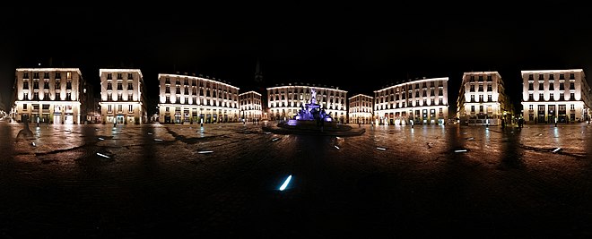 La place, de nuit, déserte, les bâtiments et la fontaine sont éclairés