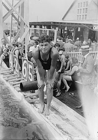 A young male swimmer stands at the edge of a pool with other swimmers sitting in the background. He leans over and squints into the camera. He wears circa 1930 swimming attire with a tank top.