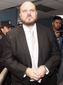 An image of Alejandro Muyshondt in handcuffs standing and wearing a business suit and tie being followed by a police officer and a news reporter