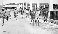 Black and white photograph of people on a road. Three men on the right hand side of the photo are carrying rifles.