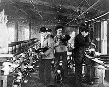 Four men working in a rustically equipped laboratory. Benches covered with equipment are on both sides. On bench is supported by a wooden barrel. Wires can be seen suspended from the ceiling.