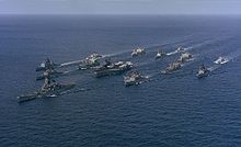 A large collection of ships sailing on the sea from the back right to the front left. At the center of the cluster of ships is an aircraft carrier, with a battleship in front of the carrier. Other ships of various types are sailing in a roughly circular formation to provide defense for the aircraft carrier.