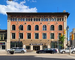 The front of a four storey building. On the main floor, the facade is brown tile. Floors above that it is terracotta-colored stucco. Just below the roofline is a detailed mosaic of many characaters represented in flat, egyptian-style design. Just below the fourth floor windows are decorative circles containing more mosaics. The over all design is very busy and detailed, while being symmetrical and solid.