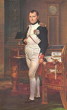 Full length portrait of Napoleon in his forties, in high-ranking white and dark blue military dress uniform. He stands amid rich 18th century furniture laden with papers, and gazes at the viewer. His hair is Brutus style, cropped close but with a short fringe in front, and his right hand is tucked in his waistcoat.