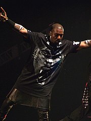 A man holding a microphone in his right hand whilst on stage. He wears a black T-shirt with a tiger-like face on it, tight leather pants, and a kilk.