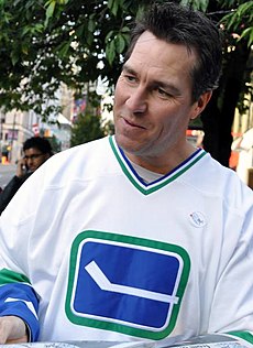 A middle-aged, Caucasian man wearing a white ice hockey jersey with a blue and green logo of a horizontal hockey stick. His head is tilted and he is looking towards the right. His hair is dark brown and spiked upwards in the front.