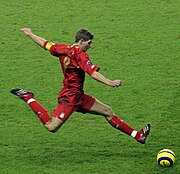 Steven Gerrard wearing a football kit, about to strike a ball. On his forearm, he is wearing an armband; the letter "C" to represent "Captain" is visible.