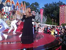 A woman wearing a long black gown. She has long golden hair and is holding a sparkling microphone. She is standing on a large red stage, surrounded by dancers in white attire. Additional background scenery include the audience and three background singers wearing white ensembles and standing on a large platform.