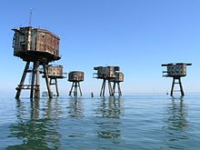 The view from a boat of a site containing six sea forts. The forts have an octagonal shape, with rusty metal walls and two rows of windows. Each fort is supported by four legs jutting from the sea at an angle