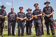 Six Calgary Police police officers stand side-by-side in a grass field. From left to right, one officer is wearing a cowboy hat and no rank insignia, one is wearing no hat and rank epaulettes revealing that she is an inspector, one is wearing a peaked cap and deputy chief rank insignia, one wears a cowboy hat and no rank insignia, one wears a peaked cap and police chief rank insignia, and another wears a cowboy hat and sergeant stripes on his sleeve.