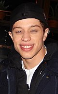 An image of a smiling Pete Davidson who is wearing a dark blue sweater and a black cap.