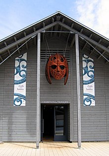 Colour photograph of the exhibition and treasury hall at the Sutton Hoo visitor centre, outside of which hangs the sculpture Sutton Hoo Helmet by Rick Kirby