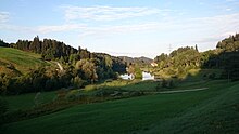 Ansicht des Wenigerweiers von Osten in Richtung Westen. Im Vordergrund ist eine Naturwiese mit einem Feuchtgebiet zu sehen, dass an den Weiher angrenzt, in dem auf einem kleinen Inselchen ein kleiner Baumbestand wächst. Der Weiher ist zwischen zwei sanft ansteigende Hängen eingebettet.