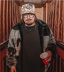 A waist-up view of singer Hardy, wearing a cap, black shirt, and jacket, and holding a red cup.
