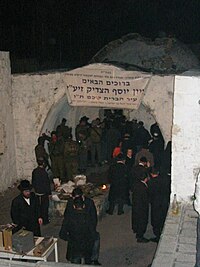 A colour photograph showing a cluster of black-garbed men in hats, along with a cluster of uniformed soldiers, standing under a whitewashed, arched opening with a doorway behind and a low dome dimly visible in the background