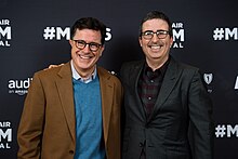 Photo of Oliver, wearing a dark suit, standing next to Colbert, wearing a tan suit, both laughing and standing in front of a press backdrop.