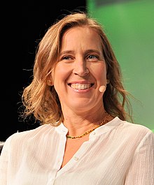 Wojcicki speaking at a TechCrunch panel. She is a middle-aged woman with blonde hair, wearing a white formal shirt and a golden necklace.