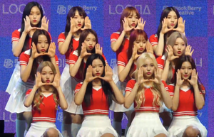Twelve young women all wearing identical red short sleeved knitted crop tops and white tennis skirts standing in a 4 by 3 configuration with their hands up to their faces in a 'shouting' gesture. The four girls in the top row are standing straight, while the four girls in the middle row are leaning forward slightly, and the four girls in the bottom row are kneeling on one knee.