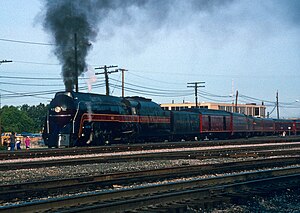 A black streamlined steam locomotive with a 4-8-4 wheel arrangement (four leading wheels, eight driving wheels, and four trailing wheels) and its tender, pulling an extra tender, a Tuscan red baggage car, and a matching set of Tuscan red passenger cars