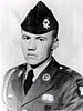 Head and shoulders of a young white man wearing a garrison cap with a large badge on the left side and a military jacket with a braided shoulder cord and several patches and pins on the lapels, shoulders, and chest.