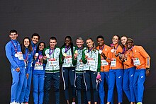 Photo of a row of twelve athletes in front of a dark grey background, four representing Italy with bronze medals wearing blue track suits, four representing Ireland with gold medals wearing green and blue track suits, and four representing the Netherlands with silver medals wearing orange and blue track suits