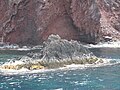 A detail of the west coast shows the reddish volcanic rock cliffs and a small rock islet with a visible intertidal algal zone