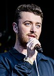 Sam Smith, a White transmasculine individual with a beard, holding a microphone to their mouth and singing.