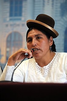 A woman wearing a white top and a small, crooked bowler hat speaks into a microphone. These bowler hats have been worn by Quechua women in this way since the 1920s.