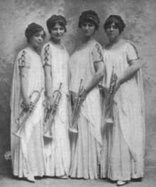 Four young white women standing in a row, wearing matching white flowing classical-style gowns, all holding trumpets