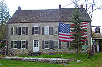 A large stone house with a large American flag draped across its upper right story behind an evergreen tree. In front of it is a blue and gold historical marker.