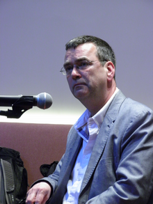 Mick Herron, wearing a suit, sitting at a Microphone.