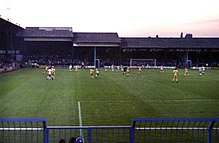 A professional football match in progress, viewed from behind one of the goals. One team is in white and the other is in yellow.
