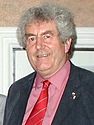 A photograph of a smiling man with grey hair wearing a black suit with a pink dress shirt and a red tie
