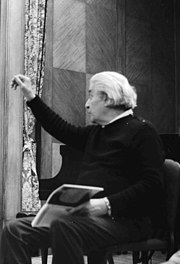 Photographie en noir et blanc d'un homme assis aux cheveux blancs, ayant le le bras droit levé.