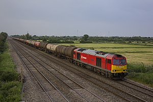 a locomotive, in DB Schenker Rail livery (red with a light grey roof and yellow front panel), hauling wagons on a four-track section of railway, past green fields