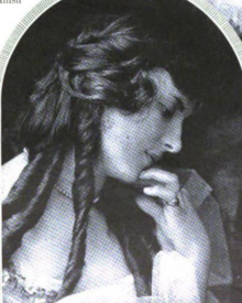 A young white woman with dark hair set in long curls; one hand on her chin; photographed in profile